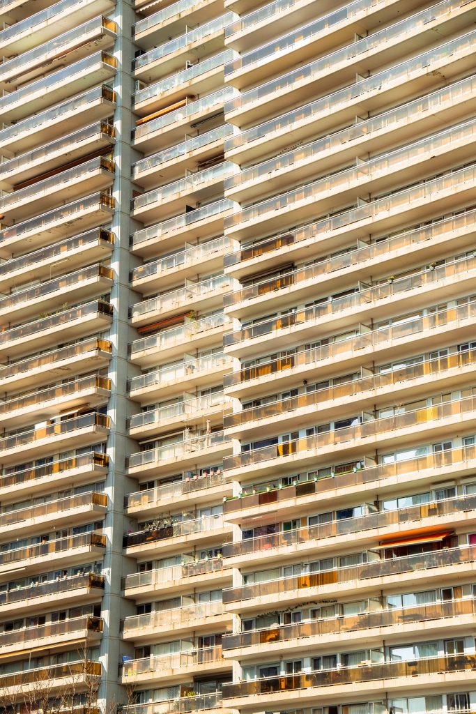 Flat-on close-up of a tall Brussels residential apartment tower with repeating rows of balconies and glass railings bathed in warm late-afternoon golden light