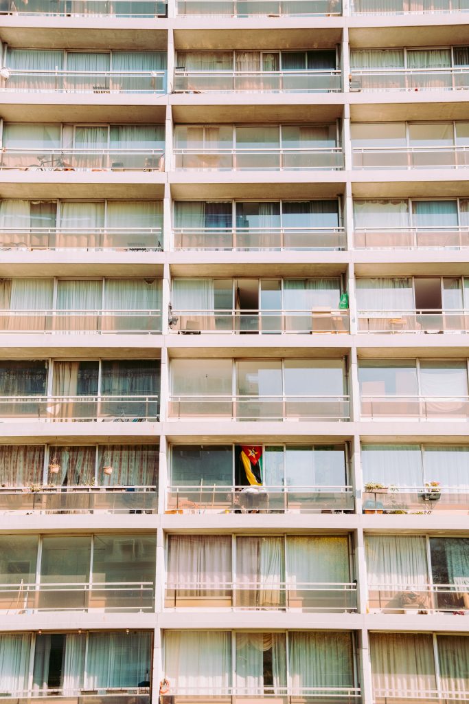 Close-up of a large Brussels residential apartment block facade showing a regular grid of balconies with sheer curtains, a Mozambique flag visible on one balcony, warm afternoon light