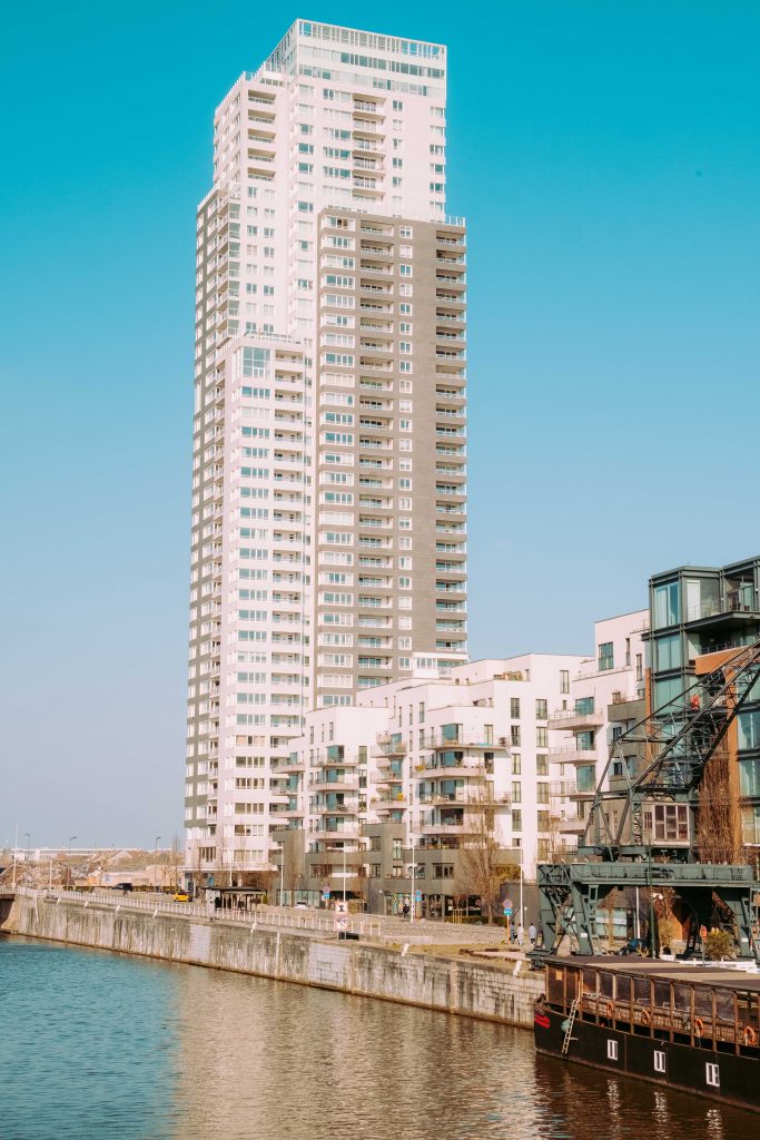 Wide view of Brussels canal waterfront in the Tour and Taxis area, tall white residential tower dominating the skyline against clear blue sky, old industrial crane and moored boat in the right foreground
