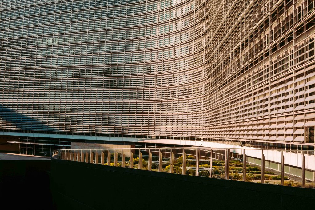Wide-angle colour photograph of the Berlaymont European Commission building in Brussels showing the curved aluminium facade in warm afternoon light, with a green planted roof garden and glass colonnade at ground level below