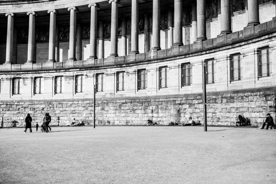 Black and white photograph of people sitting and lying against the curved stone base of the Cinquantenaire colonnades in Brussels, wide sandy plaza in foreground, ionic columns and frieze above